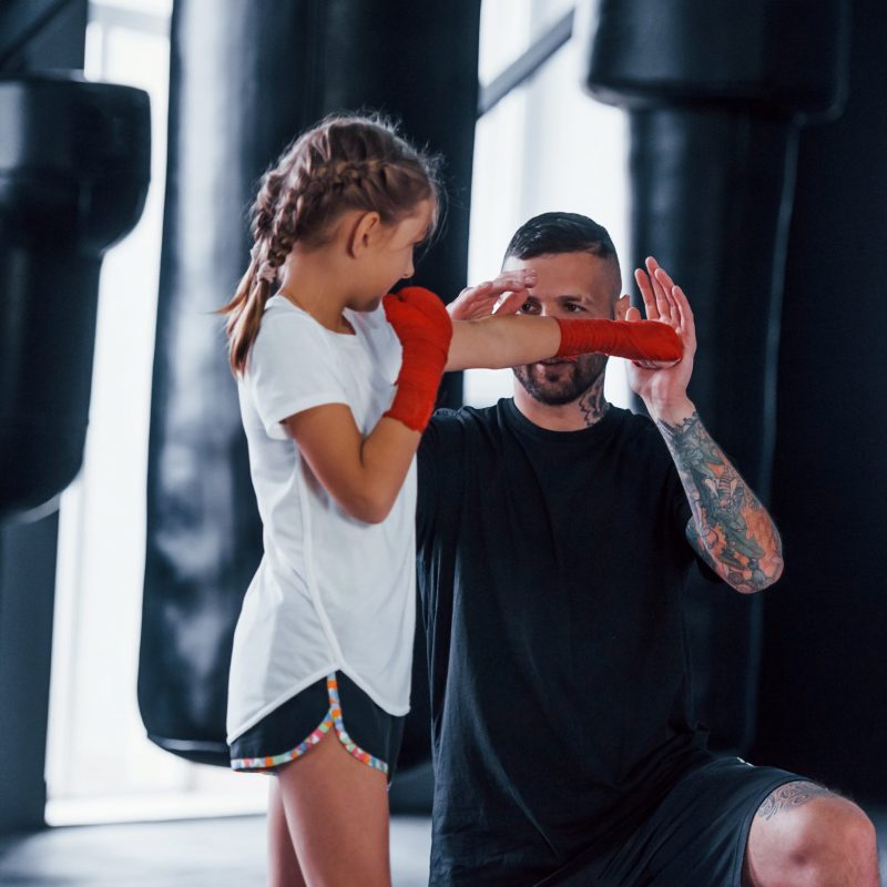 Basic punches. Young tattooed boxing coach teaches cute little girl in the gym.